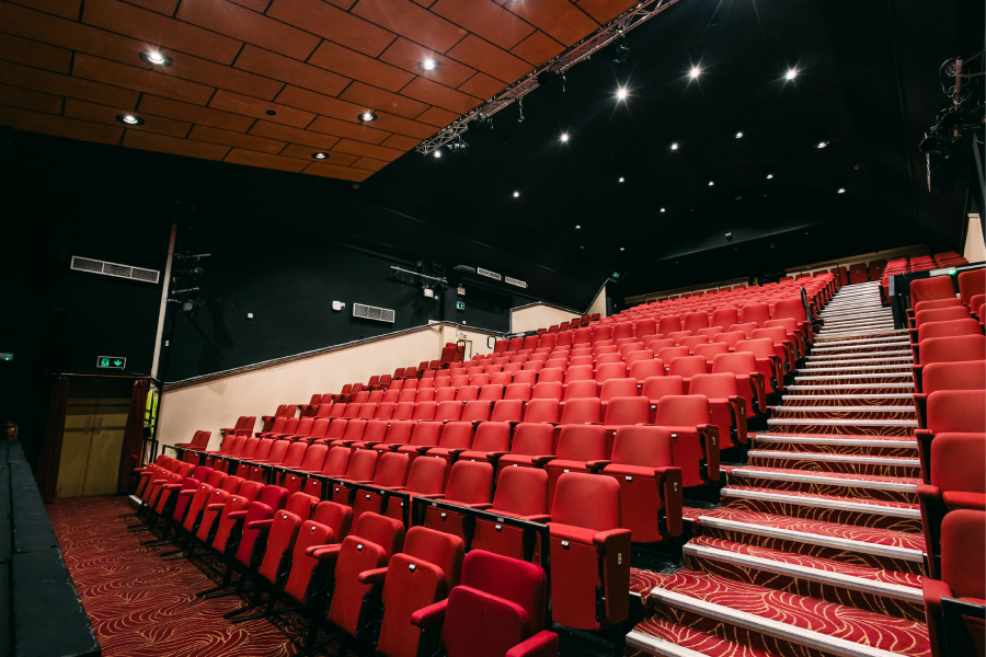 Theater with rows of vibrant red seats and patterned carpeted stairs. The dim lighting and wood-paneled ceiling create a warm, inviting atmosphere.