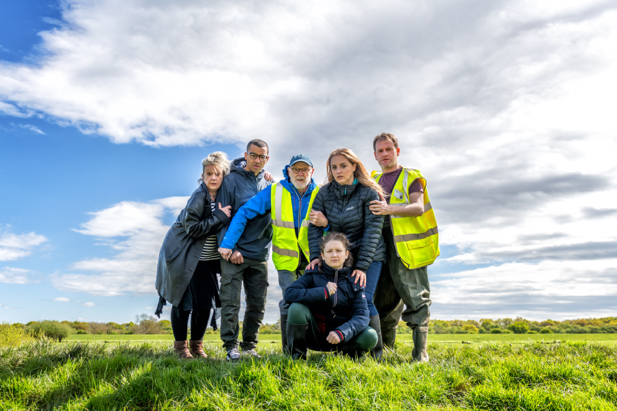 A group of six people, wearing jackets and high-visibility vests, stands closely on a grassy landscape under a cloudy sky, conveying teamwork and determination.